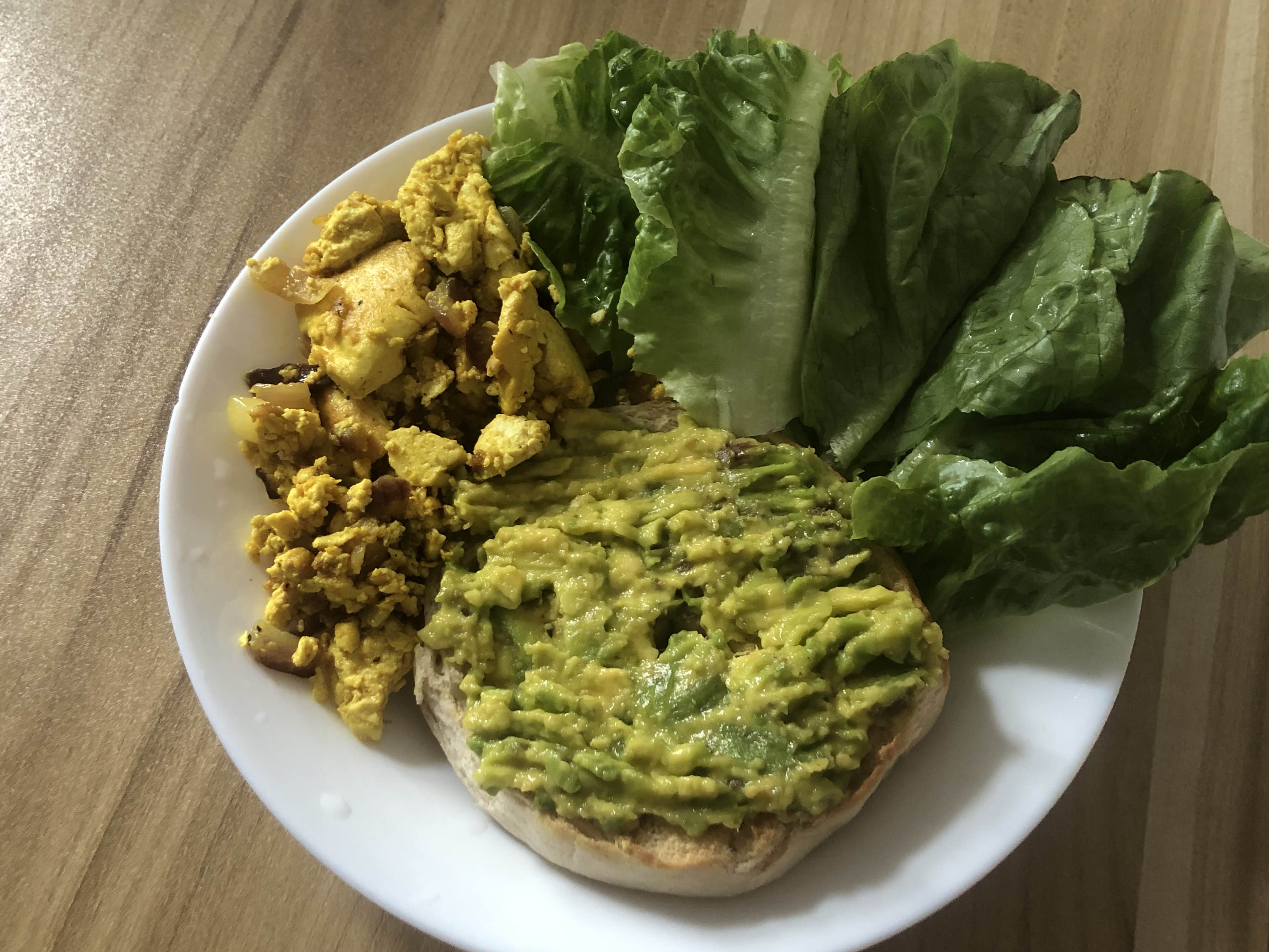a plate of food containing a potion of tofu scramble, mashed avocados spread on a half of a bagel, and portion of lettuce.